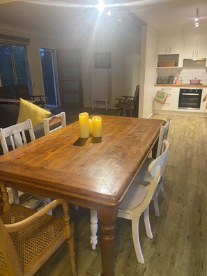Un comedor con una mesa de madera, sillas y velas. Se observa una cocina al fondo con armarios y electrodomésticos.