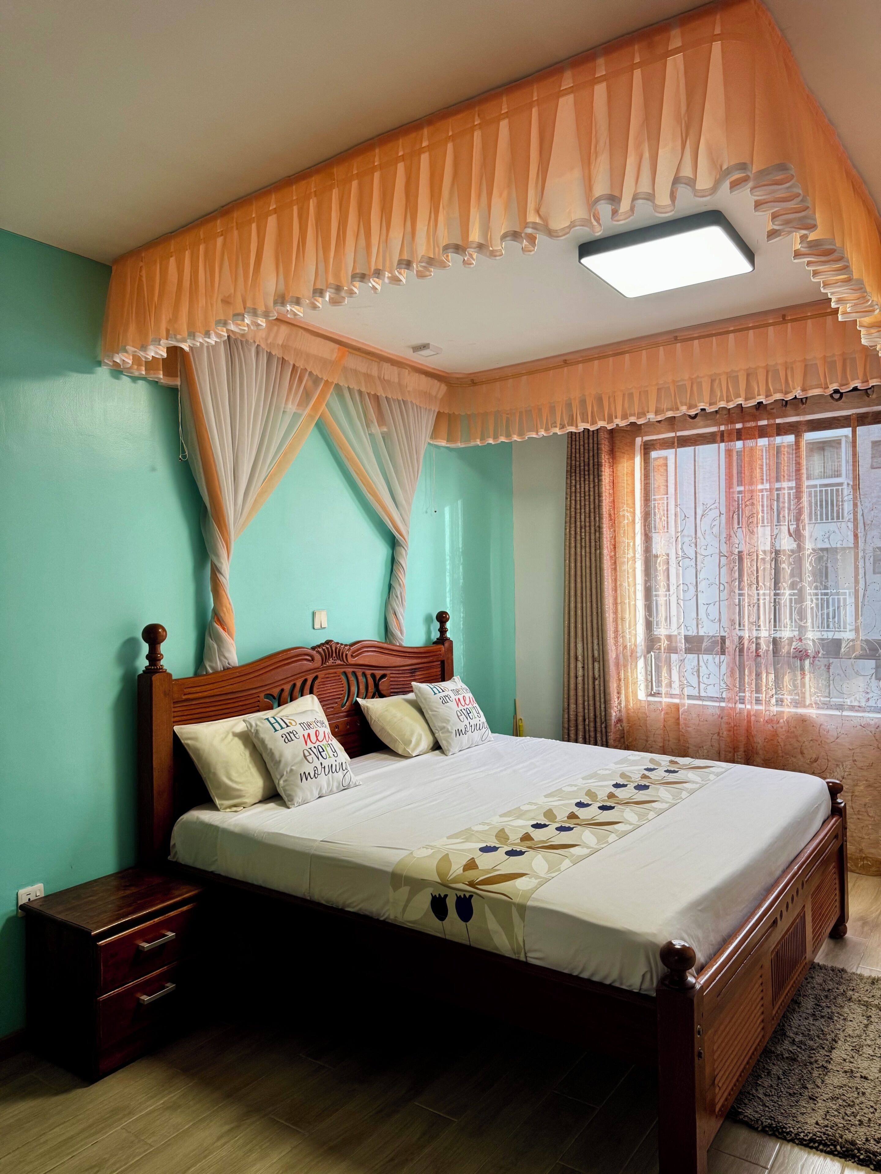 A bedroom with a bed, wooden headboard, bedside tables, a window with curtains, and a ceiling light.