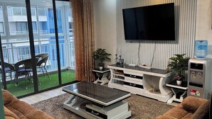 Un salon moderne doté d’un téléviseur à écran plat, d’un lustre, d’un coin repas avec des chaises et d’une grande fenêtre donnant sur un balcon.
