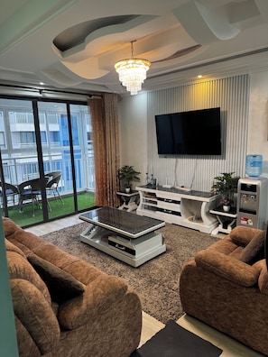 A modern living room with a flat-screen TV, a chandelier, a dining area with chairs, and a large window with a view of a balcony. - Schnell Homes (Syokimau)