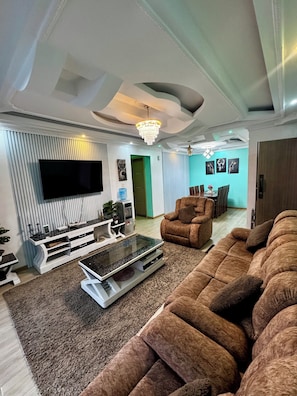 A modern living room with a large flat-screen TV, a brown sofa, a coffee table, and a chandelier. - Schnell Homes (Syokimau)