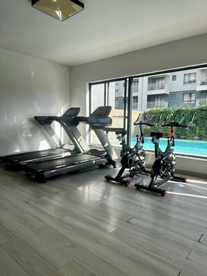 A gym with treadmills and an elliptical machine, large windows, and a view of a building with balconies. - Schnell Homes (Syokimau)