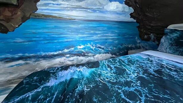 Habitació amb un mural d'una escena oceànica blava, un llit amb una colcha blava i blanca amb dibuixos, i una paret de pedra.