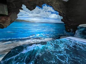 A room with a mural of a blue ocean scene, a bed with a blue and white patterned bedspread, and a stone wall.