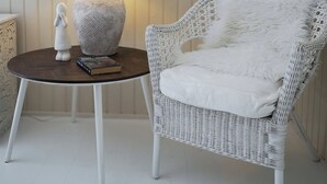 Ein weißer Korbstuhl mit einem flauschigen weißen Kissen, ein runder Holztisch mit einer Lampe und eine an der Wand montierte Uhr.