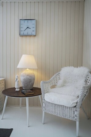 Ein weißer Korbstuhl mit einem flauschigen weißen Kissen, ein runder Holztisch mit einer Lampe und eine an der Wand montierte Uhr.