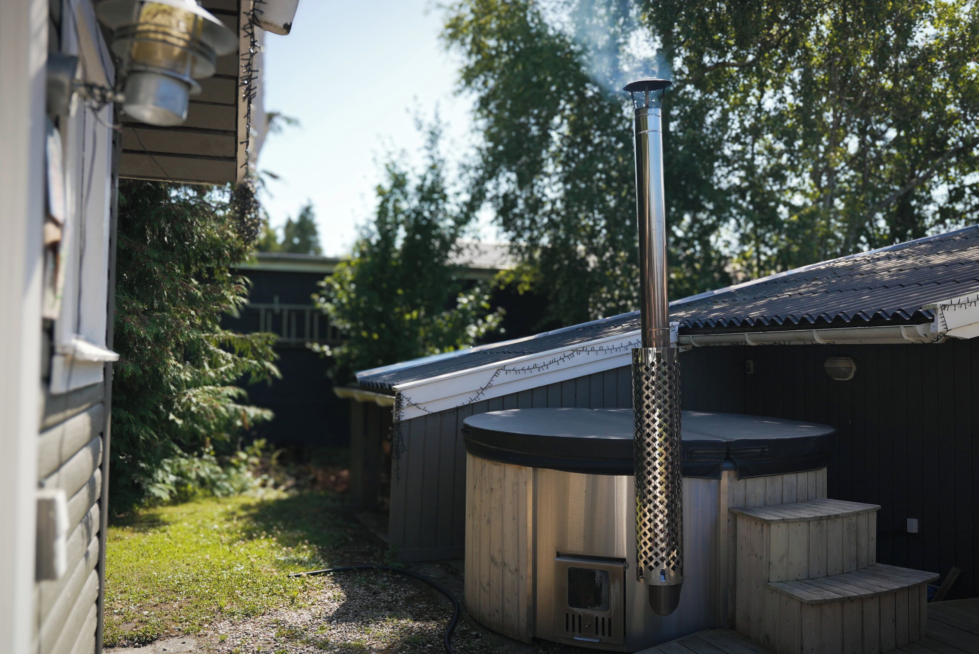 Eine Sauna mit Schornstein, aus dem Rauch aufsteigt, umgeben von Holzbauten und einem Garten.