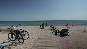 Ein Strand mit einer Holzpromenade, die zum Wasser führt, abstellbare Fahrräder und Menschen, die den Strand genießen.