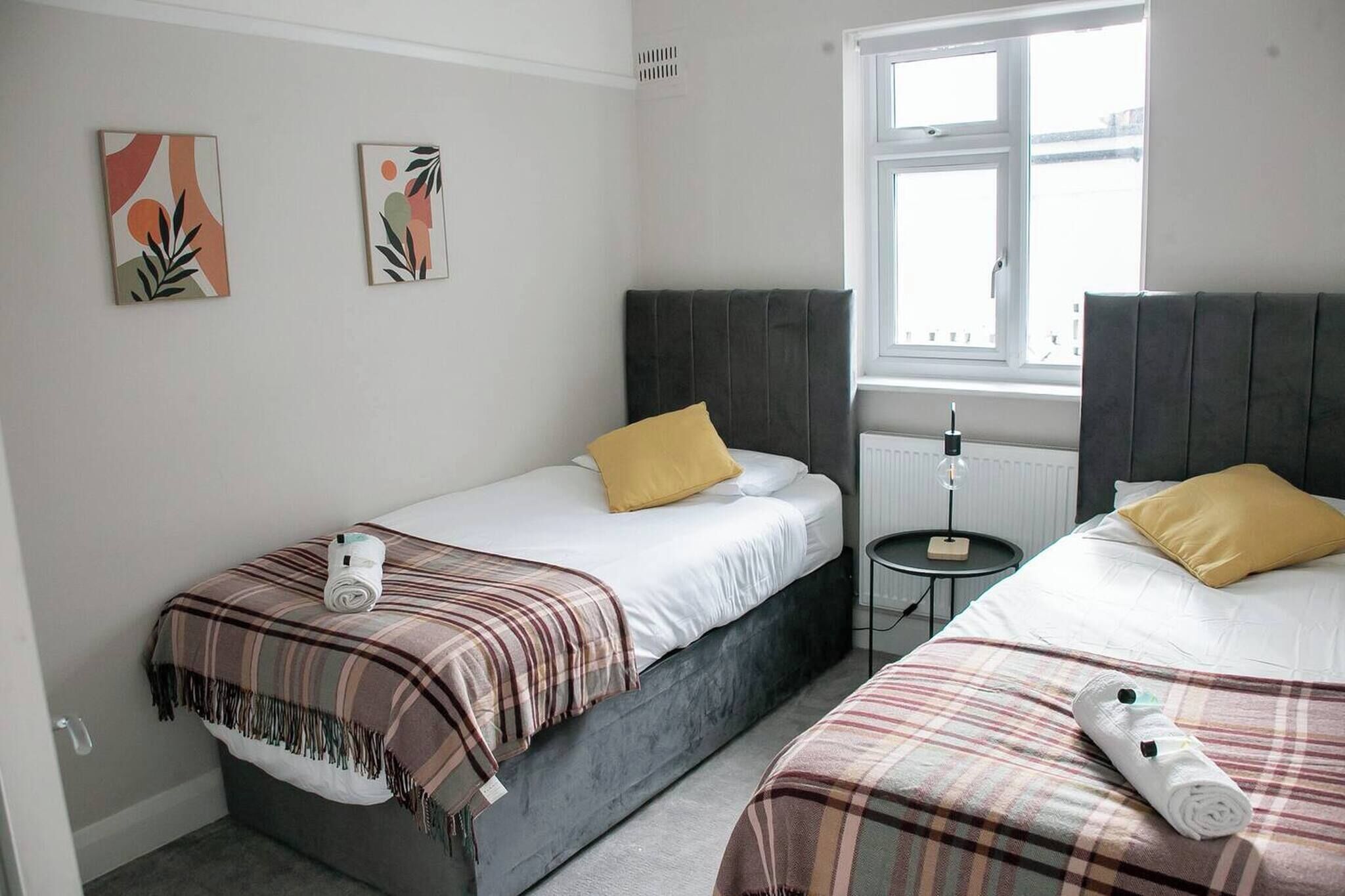 A hotel room with two beds, each with a pillow and a rolled towel. There are two wall-mounted artworks, a window with blinds, and a small table with a lamp.