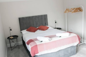 A neatly made bed with a grey headboard, a bedside table with a lamp, and a wooden hanger rack against the wall. - Art Deco Stylish 3 Bed Apartment (Leigh-on-Sea)