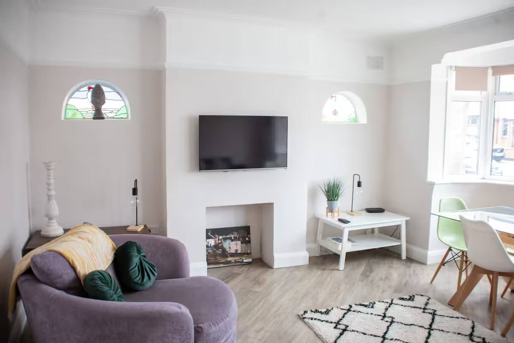 A modern living room with a flat-screen TV, a fireplace, a dining area with a table and chairs, and two windows with arched tops.