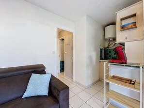Ein Wohnzimmer mit einem braunen Sofa und einem hellblauen Kissen, eine Küchenzeile mit einer roten Kaffeemaschine und ein Flur, der in einen weiteren Raum führt.