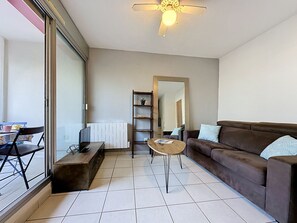 A living room with a brown sofa, a coffee table, a TV on a wooden stand, a bookshelf, and a radiator. - Les Grenadines by Interhome (La Grande Motte)