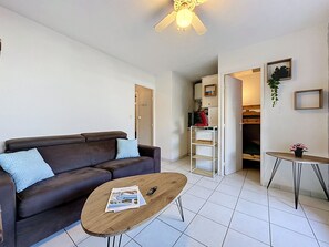 Een woonkamer met een bruine bank, een houten salontafel, een keukenhoek met een rode apparatuur en een klein tafeltje met een plant.