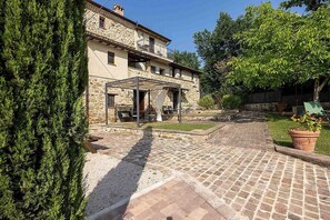A stone building with a covered outdoor seating area, a paved courtyard, and a large tree to the left. - Villa Pietrarosa: A characteristic and welcoming three-story age-old estate made of stone, with Free WI-FI. (Assisi)