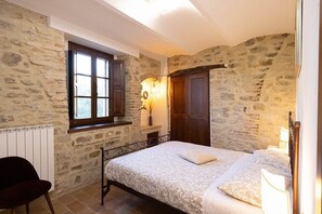 A bedroom with stone walls, a bed with a patterned bedspread, a window with shutters, and a radiator. - Villa Pietrarosa: A characteristic and welcoming three-story age-old estate made of stone, with Free WI-FI. (Assisi)