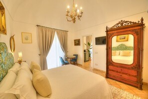 A bedroom with a large bed, a wooden dresser with a mirror, a chandelier, a desk with a chair, and a window with curtains. - Casa Minera: An elegant independent house situated at a short distance from the beach, with Free WI-FI. (Minori)