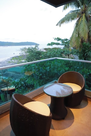 A balcony with wicker furniture, a glass railing, and a view of the sea and greenery. - Sea Deck (Port Blair)