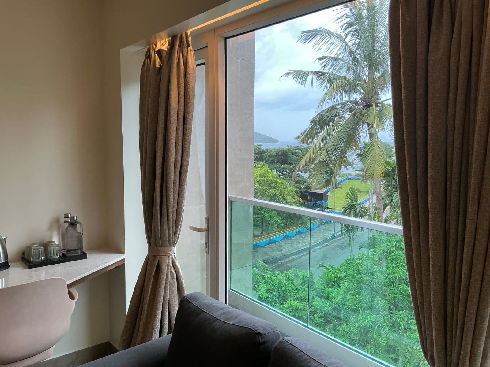 A room with a large window, a sofa, a small table with a kettle, and a view of a green landscape with palm trees.