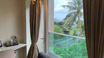 A room with a large window, a sofa, a small table with a kettle, and a view of a green landscape with palm trees.