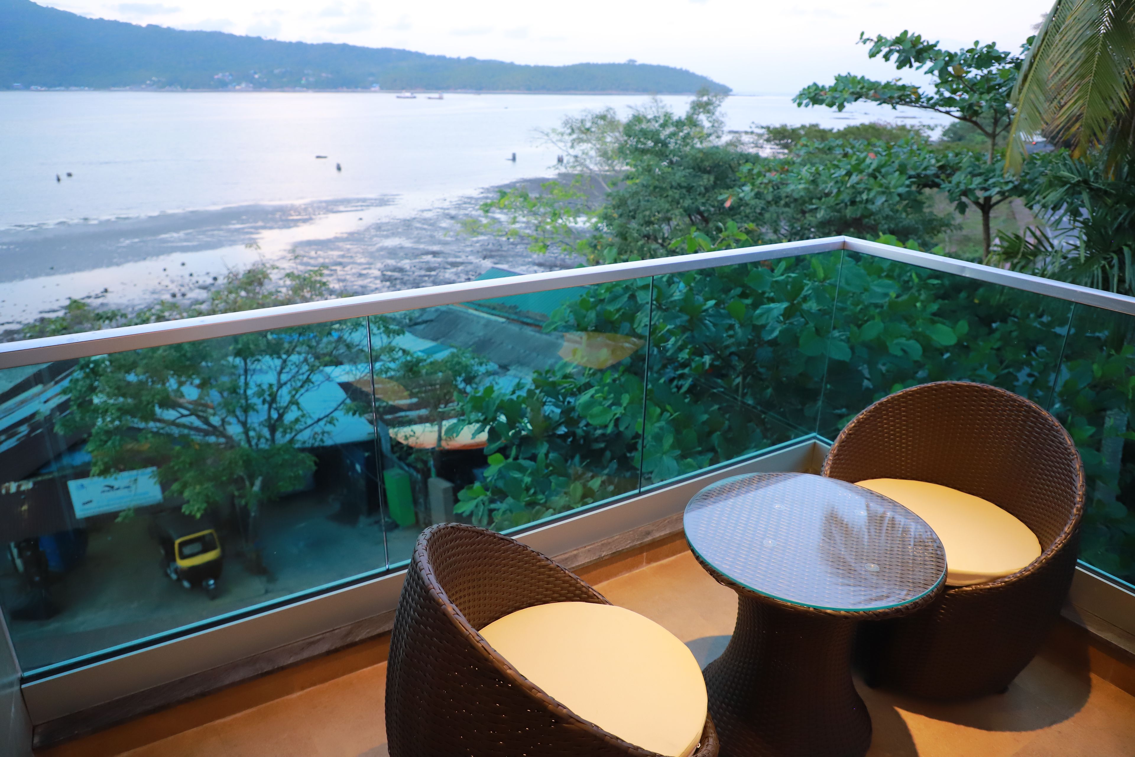 Un balcon avec des fauteuils en osier et une table en verre, d’où l’on peut admirer un plan d’eau et une côte lointaine.