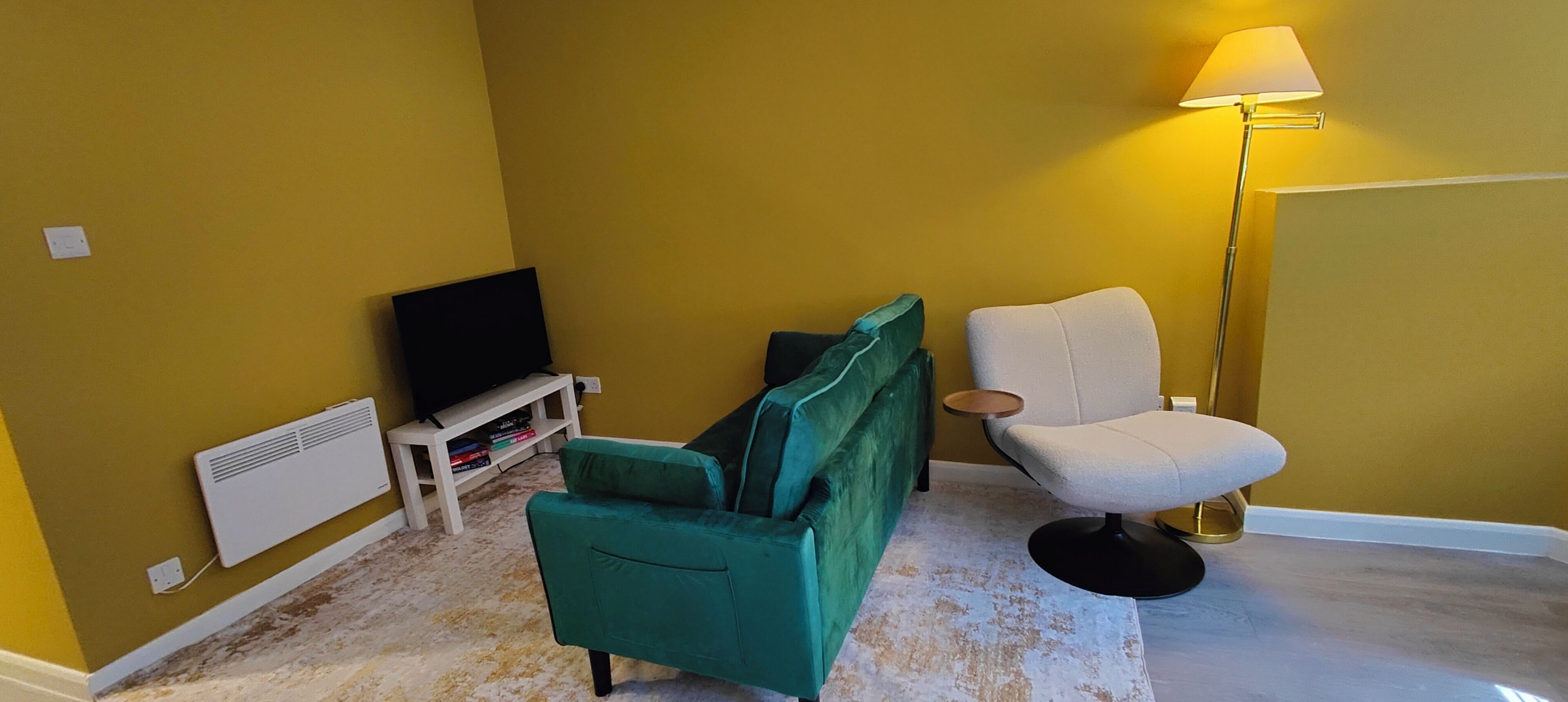 A room with a green sofa, a white armchair, a TV on a white stand, and a floor lamp.