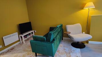 A room with a green sofa, a white armchair, a TV on a white stand, and a floor lamp.