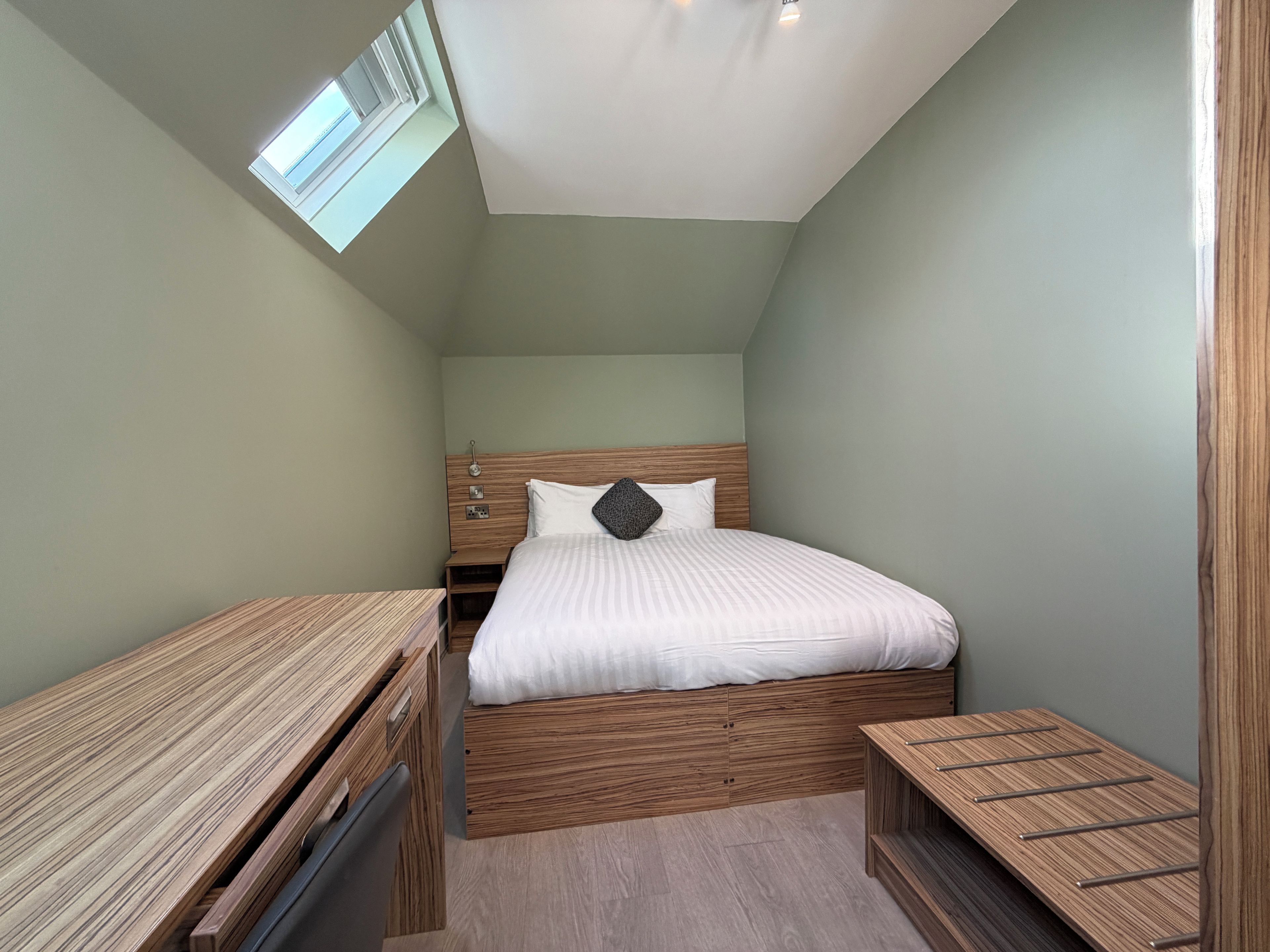 Une chambre à coucher de taille modeste, comprenant un lit simple, un bureau et une petite table, avec un plafond en pente et un puits de lumière.