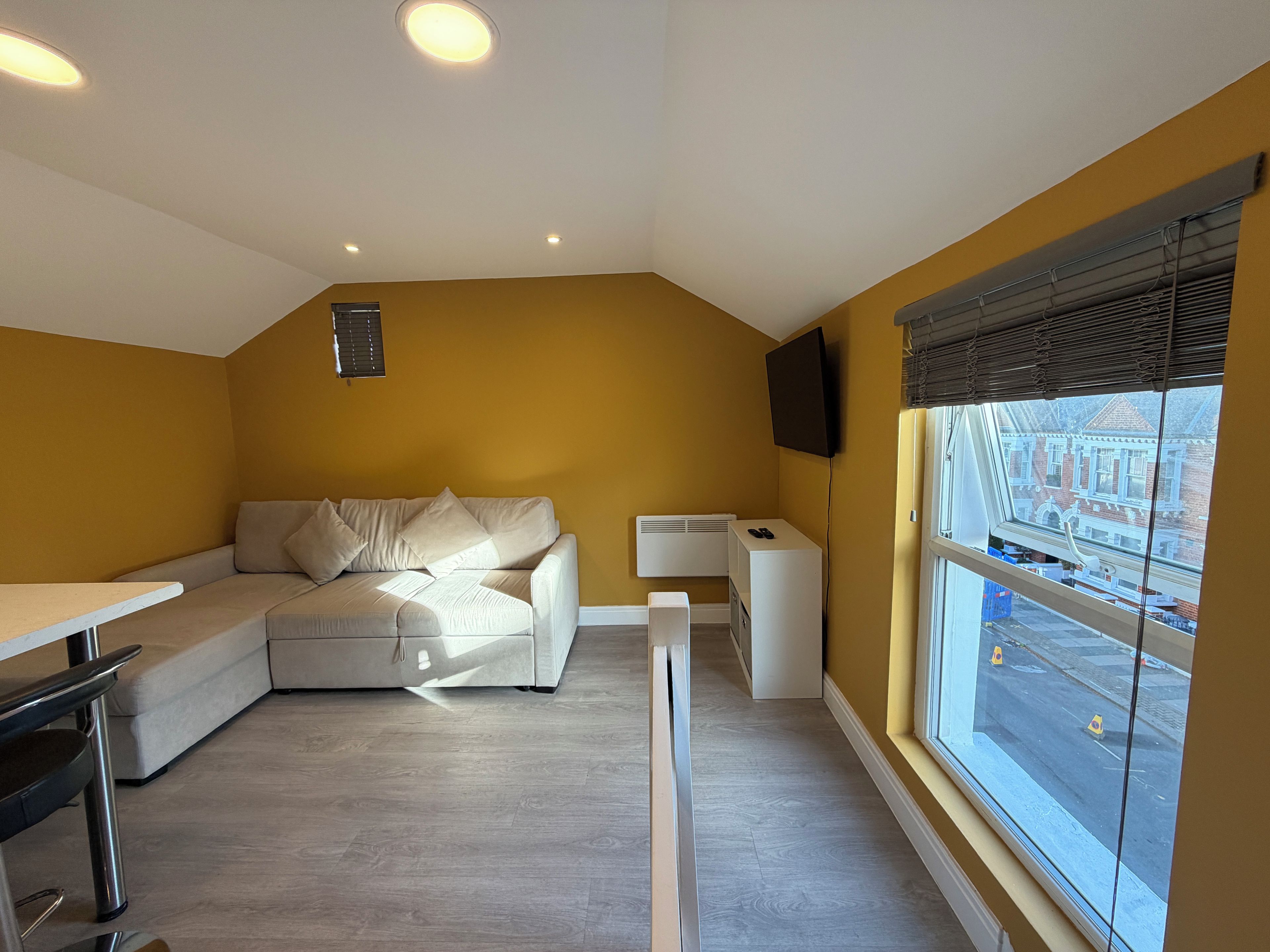 A modern living room with a sofa, a small table, a TV mounted on the wall, and a window with a view of the street.