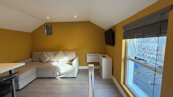 A modern living room with a sofa, a small table, a TV mounted on the wall, and a window with a view of the street.