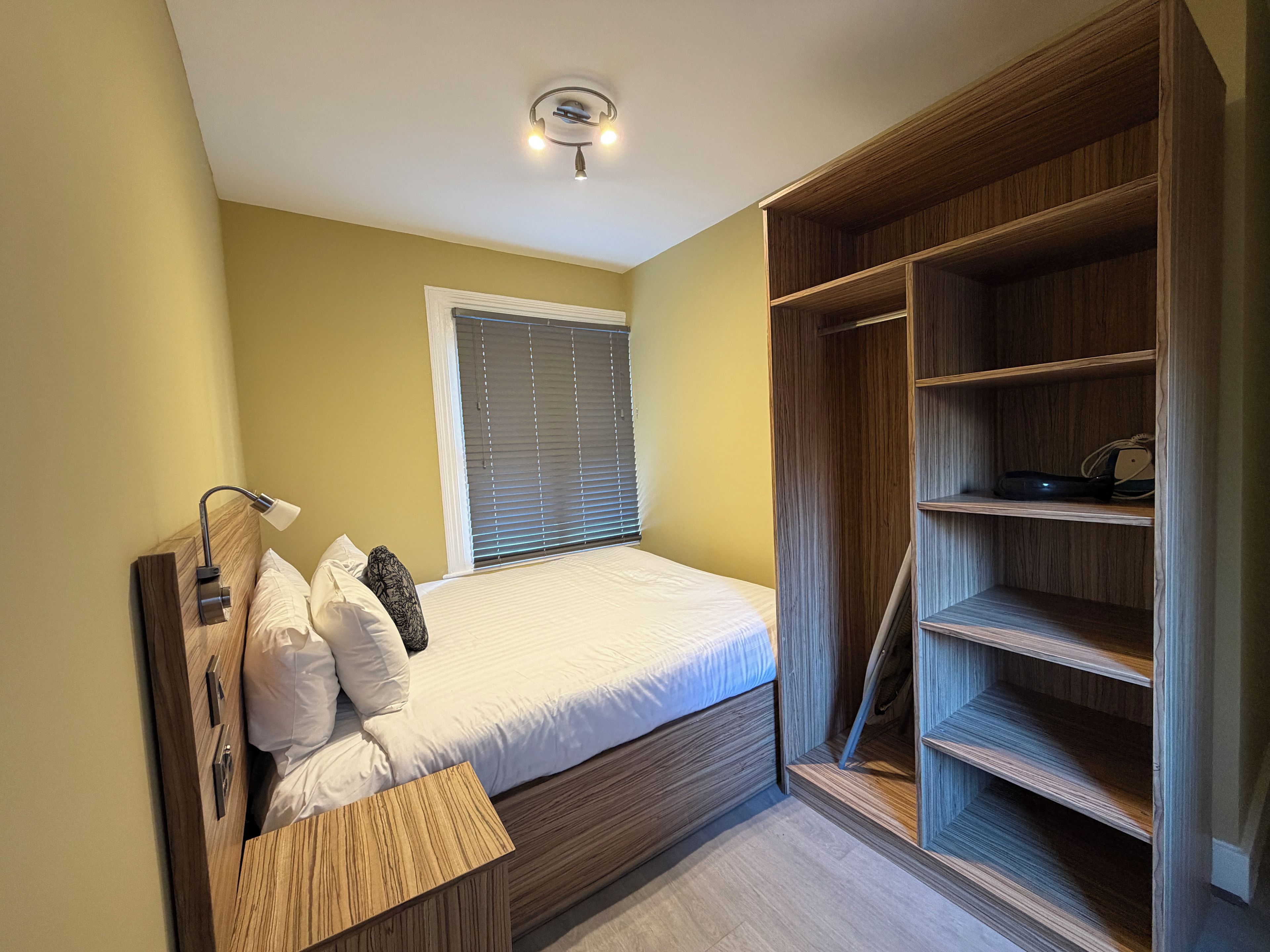 A bedroom with a bed, bedside table, lamp, and a wooden wardrobe with shelves.