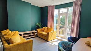 A room with teal walls, a yellow sofa, a blue armchair, and a large window with pink curtains.