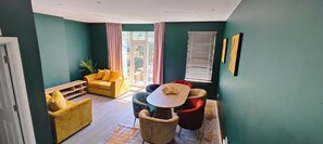 A room with a yellow sofa, a round table, and a red chair. There are two windows with curtains, a plant, and a door. - Samko Suites Clapham (London)
