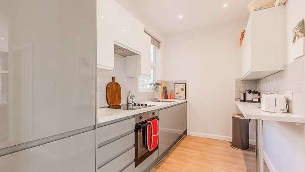 A modern kitchen with white cabinets, a wooden cutting board, a kettle, and a trash bin. - Samko Suites Clapham (London)