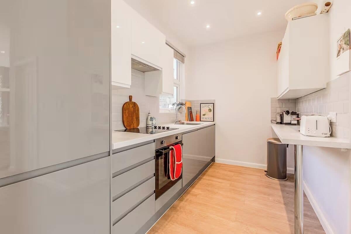 A modern kitchen with white cabinets, a wooden cutting board, a kettle, and a trash bin.