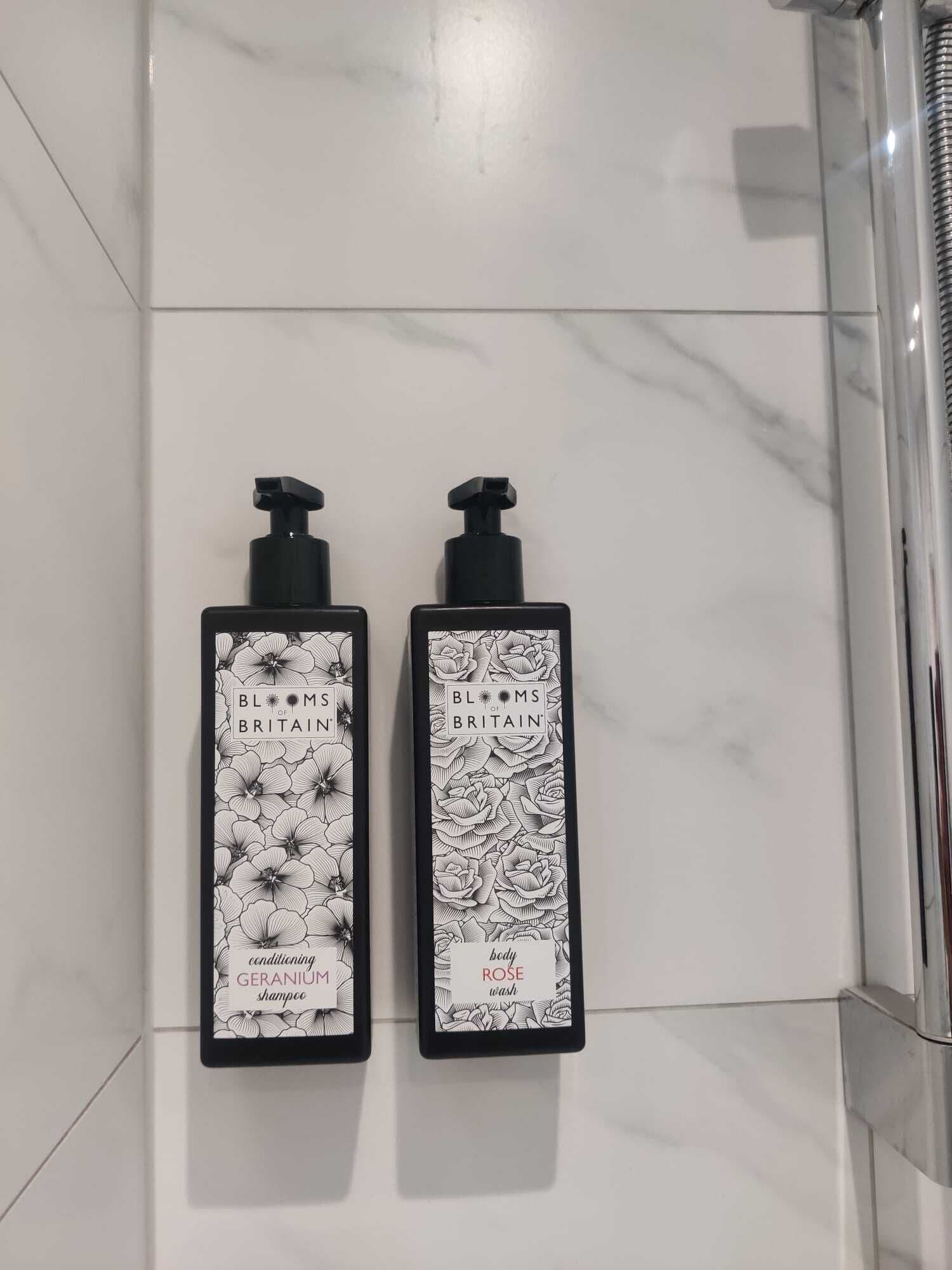 Duas garrafas pretas com padrões florais e etiquetas, uma com a inscrição 'shampoo GERÂNIO condicionador' e outra com 'gel de banho ROSA', fixadas numa parede revestida de azulejos brancos.