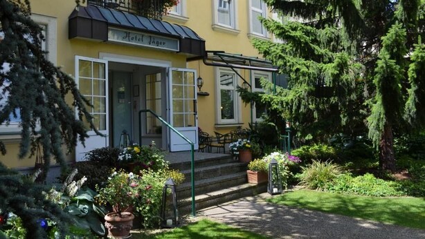 A yellow building with a sign reading 'Hotel Jäger,' a staircase, and a garden with potted plants and a tree.