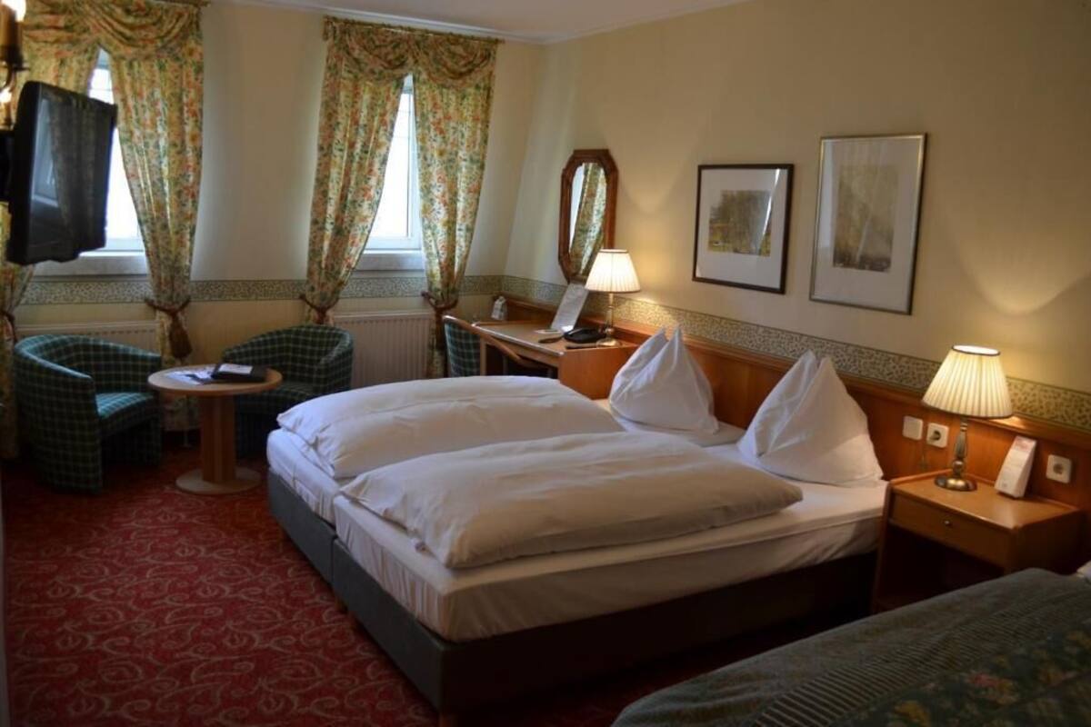 A hotel room with a bed, two chairs, a small table, a lamp, and two framed pictures on the wall.