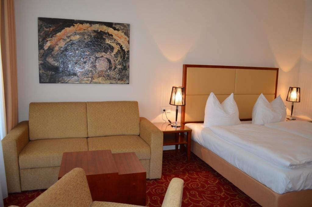 A hotel room with a sofa, a bed, a nightstand, two lamps, and a painting on the wall.