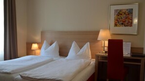 A hotel room with a bed, two bedside lamps, a desk, a chair, and a framed picture on the wall.