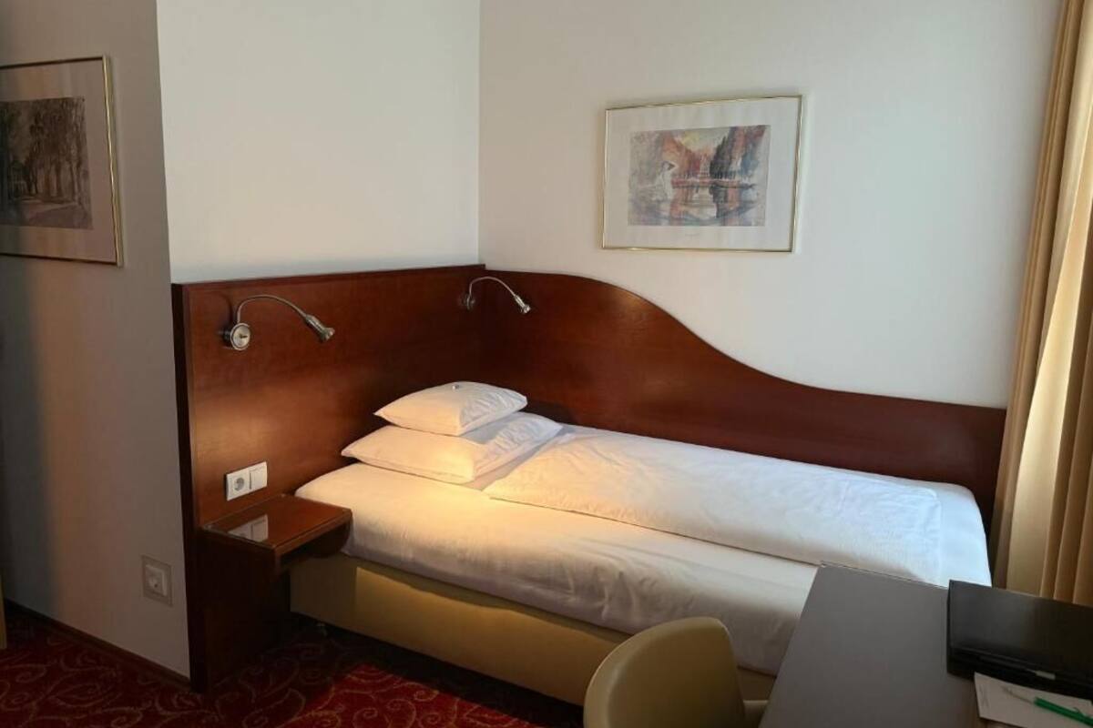 A hotel room with a single bed, a bedside table, a chair, a desk, and a painting on the wall.