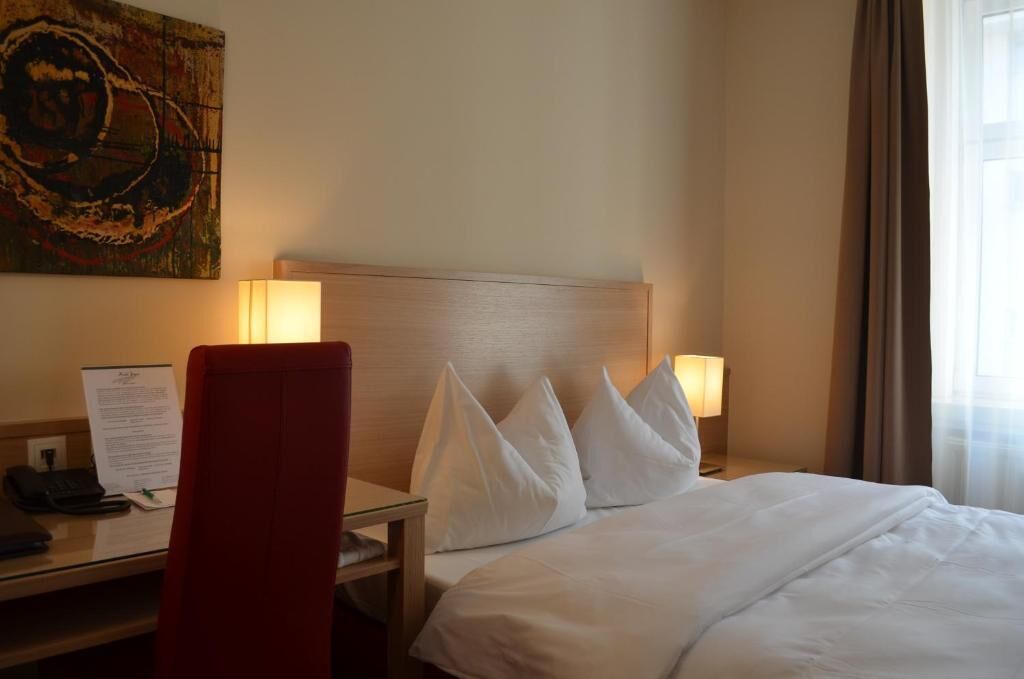 A hotel room with a bed, two bedside lamps, a desk with a chair, a telephone, and a painting on the wall.