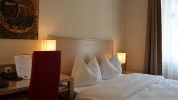 A hotel room with a bed, two bedside lamps, a desk with a chair, a telephone, and a painting on the wall.