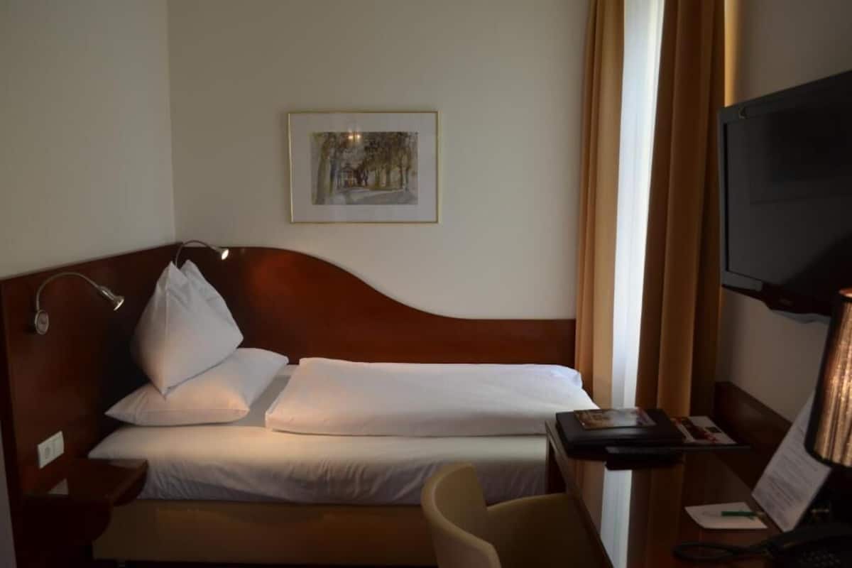 A hotel room with a bed, a desk with a lamp, a chair, a television, and a framed picture on the wall.