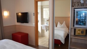 A hotel room with a bed, a red ottoman, a TV mounted on the wall, a bathroom with a sink and mirror, and a minibar. - Hotel Jäger (Wien)
