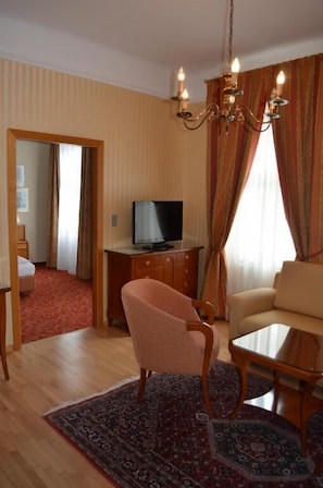 Une chambre d’hôtel équipée d’une télévision, d’un canapé, d’un fauteuil, d’une petite table, d’un lustre et d’un tapis à motifs.
