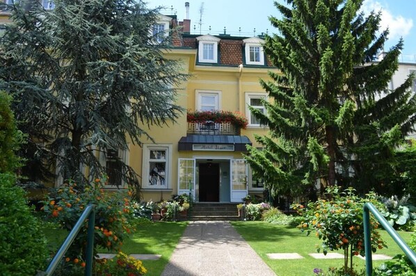 A yellow building with a balcony, a garden with flowers, and a pathway leading to the entrance.