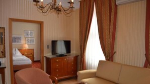 A hotel room with a bed, a television on a wooden cabinet, a sofa, a chair, and a chandelier. - Hotel Jäger (Wien)