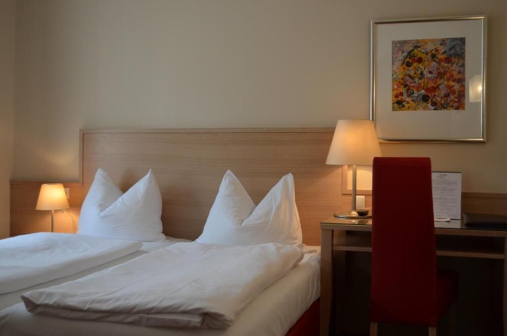 A hotel room with a bed, pillows, a desk, a chair, a lamp, and a framed picture on the wall.