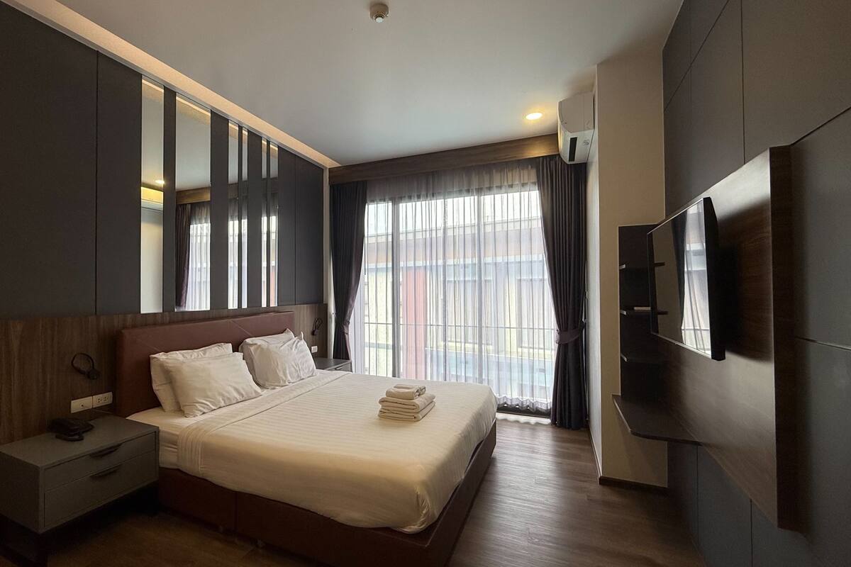 A modern hotel room with a large bed, a bedside table, a TV, and a large window with curtains.